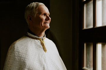 Medium closeup of modern senior Caucasian Catholic priest looking out of window, copy space