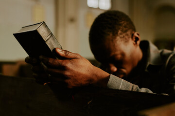 Selective focus shot of African American man sitting on bench in Catholic church holding Bible book...