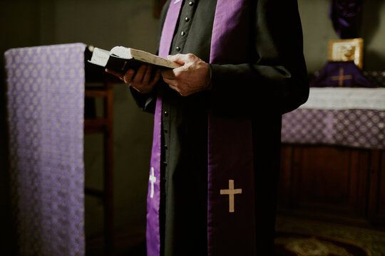 Crop shot of unrecognizable senior Catholic priest wearing soutane and stole standing indoors reading Bible