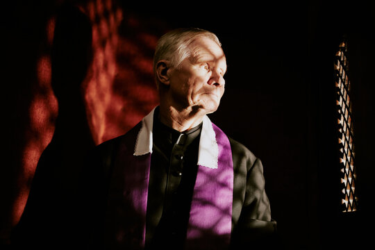 Medium closeup of senior Caucasian Catholic priest hearing out penitent in confessional booth, copy space