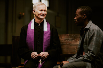 Medium shot of cheerful senior Caucasian Catholic priest talking to African American male churchgoer
