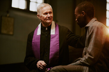 Selective focus shot of senior Caucasian priest sitting on pew supporting sad African American male...