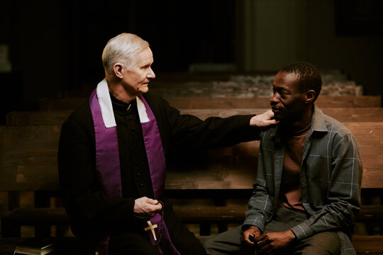 Medium shot of senior Caucasian Catholic priest and young African American man sitting on pew in church having conversation