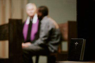 Selective focus shot on Bible book on bench in church, unrecognizable priest talking to parishioner...