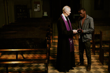 Obraz premium Long shot of ethnically diverse senior Catholic priest and young Black man standing indoors discussing religious text, copy space