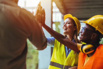 Joyful multi-ethnic construction workers in safety gear high-fiving on a site, showcasing teamwork...