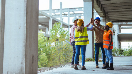A group of smiling construction workers engage in a high five on a building site, showcasing...