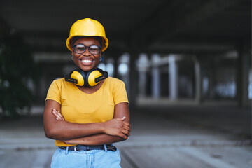 Confident black woman in yellow hard hat and safety glasses stands at a construction site, her arms...