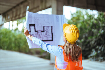 A focused female construction worker in safety gear examines architectural plans, standing at an...