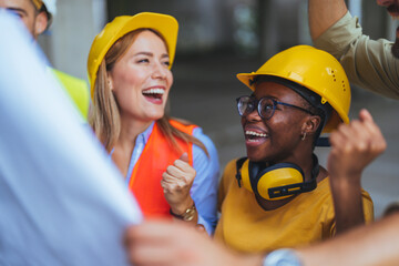 A cheerful group of diverse construction workers, in safety helmets and vests, discuss a building...