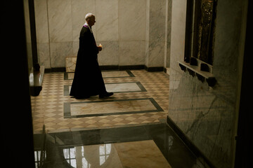 Wide shot of senior Caucasian Catholic priest holding candle walking along church corridor, copy...