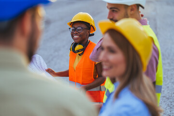 A multi-ethnic group of construction professionals in hard hats and safety vests engages in...