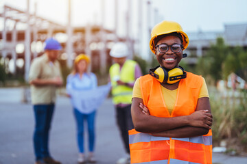 Black woman in safety gear smiles at the camera with folded arms, her team engaged in discussion...