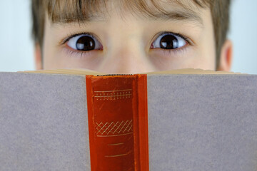 close-up of eyes of child behind thick paper book in dark red cover, learning lessons, reading...