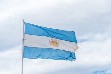 A blue and white argentinian flag with a yellow sun in the center flying in the sky.