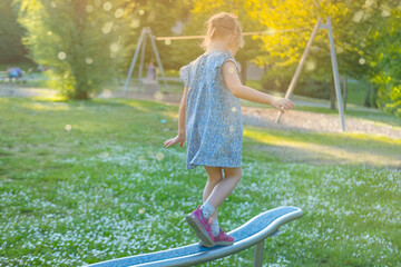 preschool girl in blue short dress walks on sports log, on playground in park, child in rays light,...