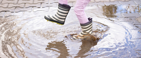 mischievous 5-year-old girl in rubber boots jumps with glee in puddle, closeup children's feet in...