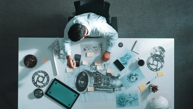 Top view of engineer looking at phone while working on tablet with green screen at meeting table with turbine engine or jet engine sketch design and various metal gear scatter around. Alimentation.