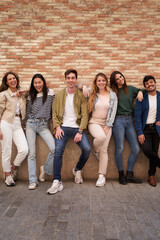 Vertical. Multiracial happy friends posing for photo looking at cheerful camera. Group portrait of smiling people leaning together on brick wall outdoor. Relationships in Generation Z and community