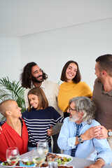 Vertical. Portrait of smiling Caucasian people looking affectionately at each other embraced together indoor at home. Multi-generational love family gathered at lunch table having fun at weekend