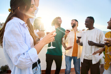 Young Caucasian beautiful woman holding wine with lens flare, multiracial friends dancing in background on rooftop. Happy generation z people having fun enjoying celebrating summer party together