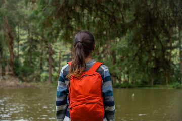 Young female traveler with a red backpack standing in front of a lake, in the background a pine forest.