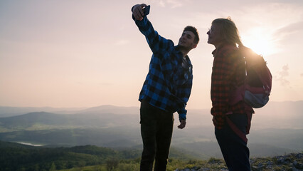 Hikers, a young woman, and a man, enjoying their success on the mountain peak, taking selfies with a mobile phone at sunset. Hiking lifestyle concept.