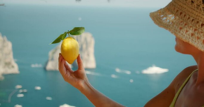 A woman in a straw hat holds a large lemon with the Faraglioni rocks and blue sea in the background from a viewpoint in Capri, Italy.