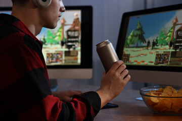 Young man with energy drink and chips playing video game at wooden desk indoors, closeup