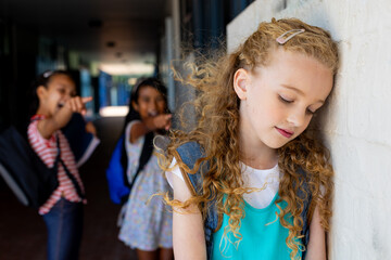 Caucasian girl leans on wall as two biracial girls whisper and engage in bull at school