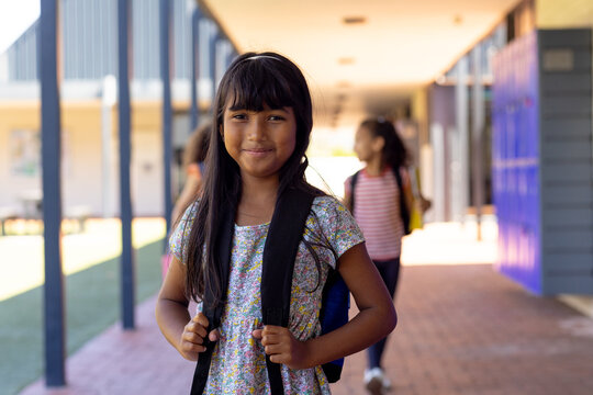 Biracial girl with long dark hair smiles in school, holding her backpack straps - Powered by Adobe