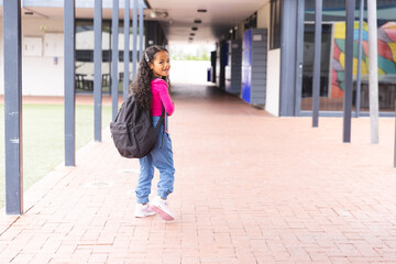 In school, young biracial girl with a backpack is walking outdoors with copy space