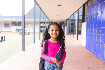 In school, young biracial female student holding a tablet stands smiling