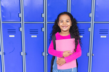 In school, young biracial girl holding a pink folder stands smiling with copy space beside lockers