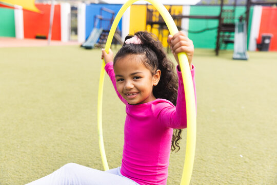 In school outdoors, biracial young girl is playing with a yellow hula hoop