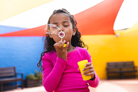 In school, young biracial girl wearing pink is blowing bubbles outdoors