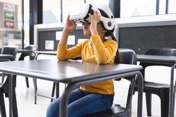 In school, young biracial girl wearing a VR headset sits at a table in the classroom