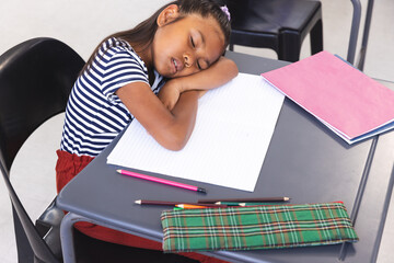 In school, young biracial girl wearing a striped shirt resting head on desk in classroom