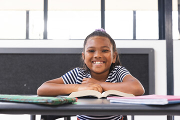In school, young biracial girl is sitting at a desk in a classroom, reading a book - Powered by Adobe