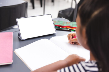 In school, biracial girl writing in a notebook in classroom, with a tablet 