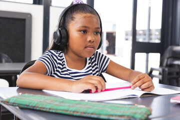 In school, young biracial girl wearing headphones focusing on reading in the classroom