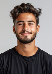 Fototapeta premium A young man with dark hair and a beard smiles softly at the camera. He is wearing a black t-shirt and standing against a plain grey background