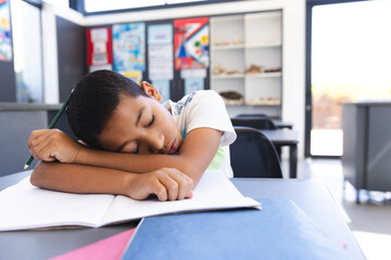 Exhausted biracial boy sleeps on desk in bright classroom.