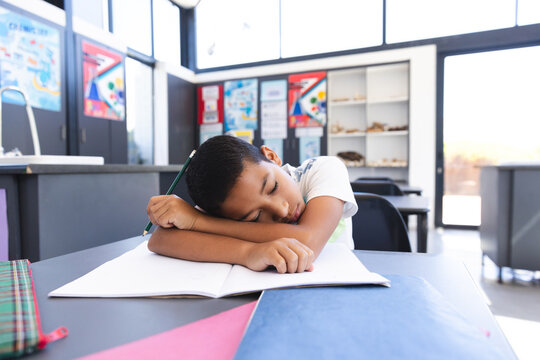 Exhausted biracial boy rests on desk during studies in bright classroom. - Powered by Adobe