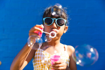 Biracial girl wearing sunglasses and a sundress enjoys blowing bubbles outdoors - Powered by Adobe