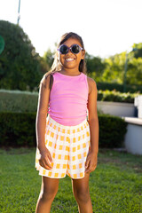 Young biracial girl in sunglasses, pink top, smiling brightly on grass outdoors