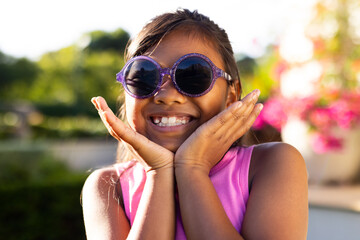 Outdoors, young biracial girl wearing sunglasses smiling with hands on cheeks