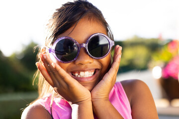 Outdoors, young biracial girl smiling brightly while wearing large, round sunglasses