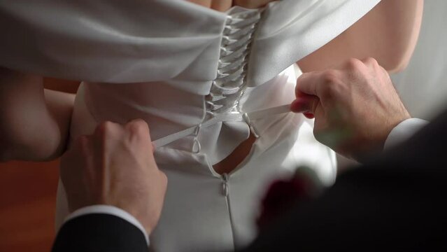 A person tying a corset on a dress. Put on wedding dress. Bride and her groom. Young woman wearing a white gown.