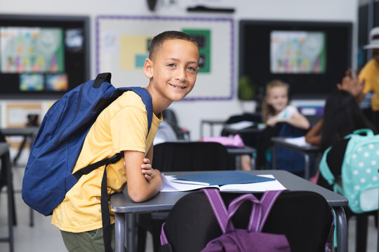 Biracial boy with a backpack stands in a school classroom, smiling, with copy space